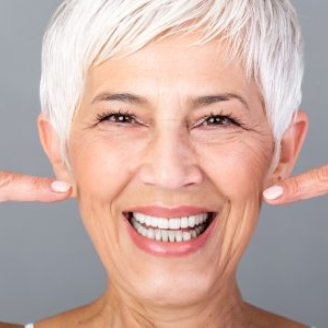 Primer plano de una mujer mayor de cabello corto y canoso, sonriendo ampliamente. Tiene las manos levantadas y apunta con ambos dedos índices hacia su boca, destacando sus dientes.